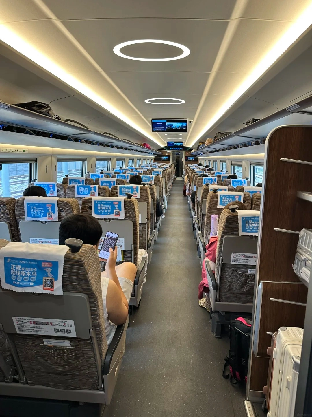 Interior view of a high-speed train carriage with rows of fabric-covered seats. The aisle runs down the center of the carriage, flanked by seats on either side. Overhead compartments are visible, as are circular lights on the ceiling. A passenger in the foreground is looking at their phone, with their legs extended into the aisle.