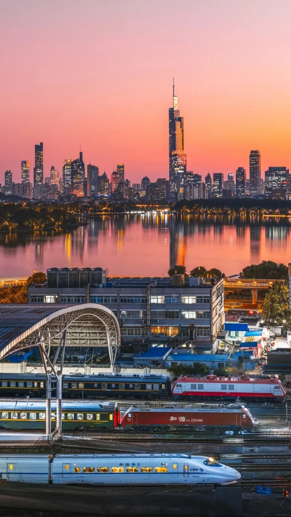 A modern white high-speed train with illuminated windows sits on train tracks in the foreground. Behind it, several other trains are visible at a station with a covered platform. In the mid-ground, a multi-story building with large windows and rooftop air conditioning units is prominent. Beyond the building, a wide river reflects the warm colors of a sunset sky. In the background, a city skyline with numerous skyscrapers, including a very tall, slender one, is silhouetted against the pink and orange sunset.