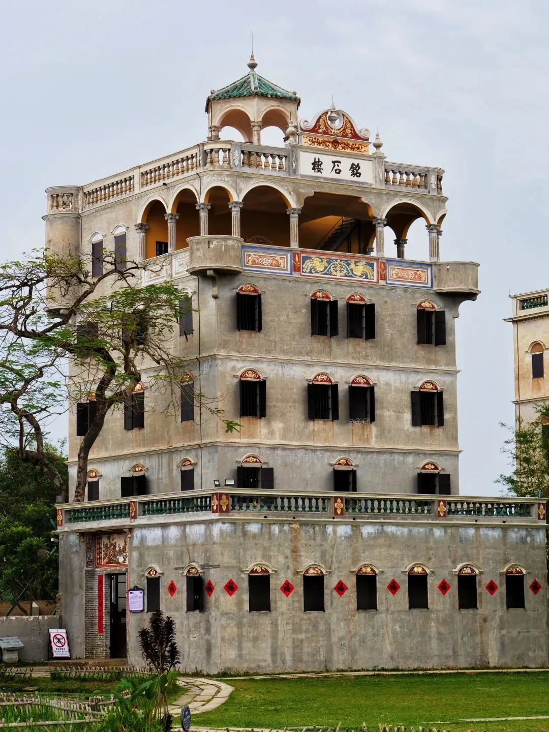 A tall, multi-story stone watchtower with a green tiled roof and balconies, surrounded by trees and green grass. The building has many windows with dark shutters and decorative elements on its upper levels.