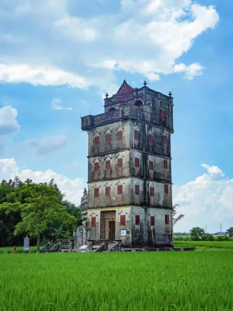 A tall, multi-story, weathered concrete watchtower with numerous small, red-shuttered windows stands in a bright green rice paddy field under a blue sky with white clouds. The tower has a distinct architectural style with balconies and a decorative roof. Trees and other less prominent structures are visible in the background.