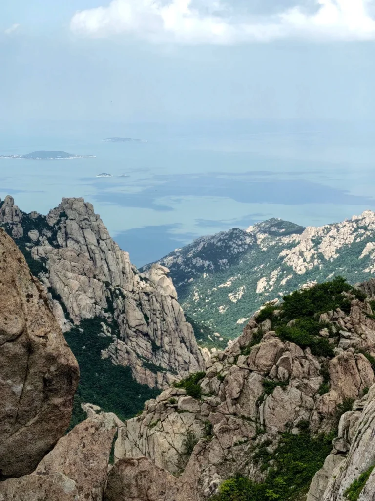 View from a high elevation on Laoshan Mountain looking out over rugged, grey granite peaks and green, tree-covered valleys towards the Yellow Sea, with several small islands visible on the horizon under a partly cloudy sky.