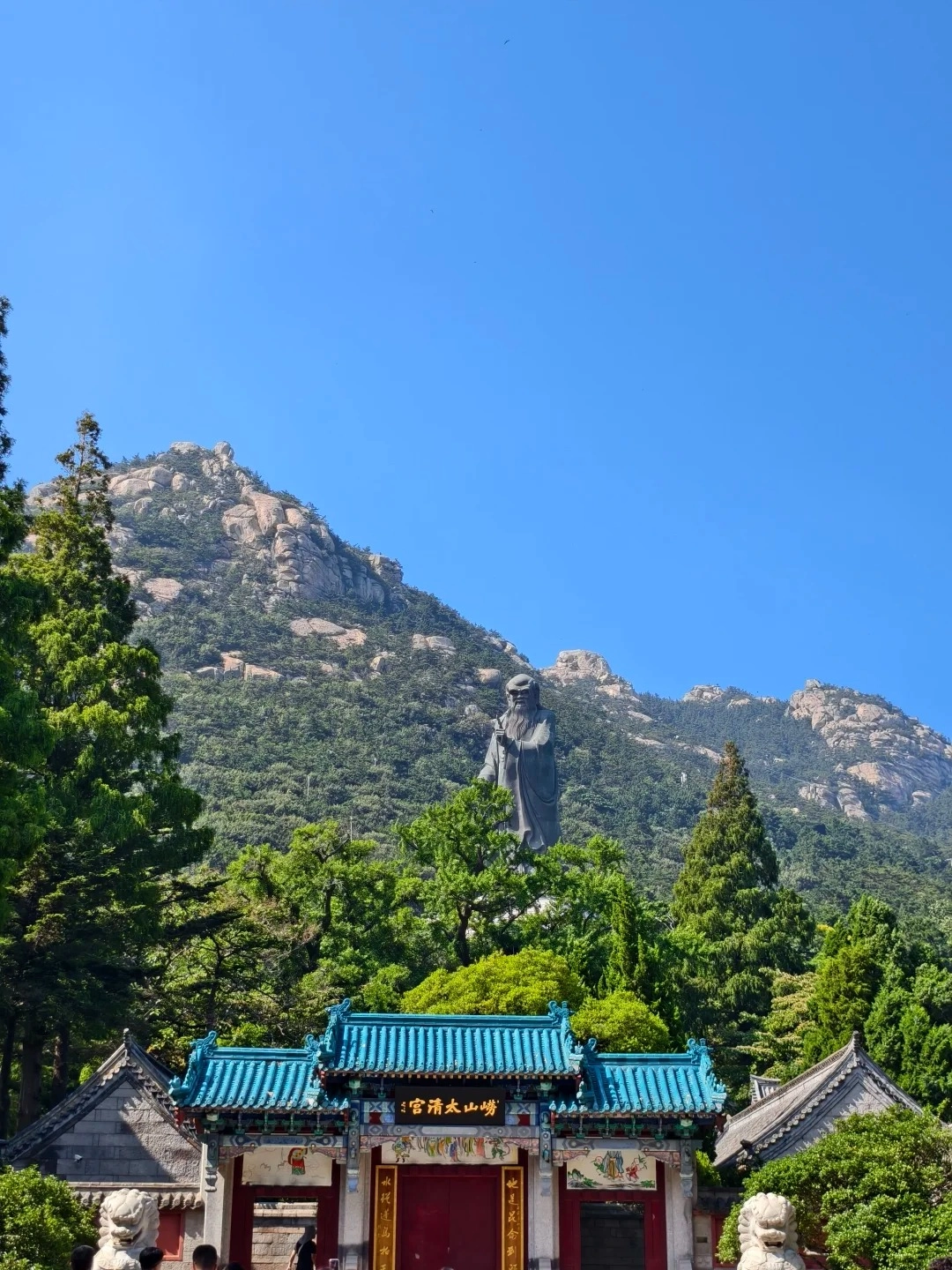 A wide-angle shot shows a majestic statue of Confucius standing on a lush green mountainside under a clear blue sky. In the foreground, a traditional Chinese palace gate with a turquoise tiled roof and red doors is visible, flanked by stone lion statues and people entering.