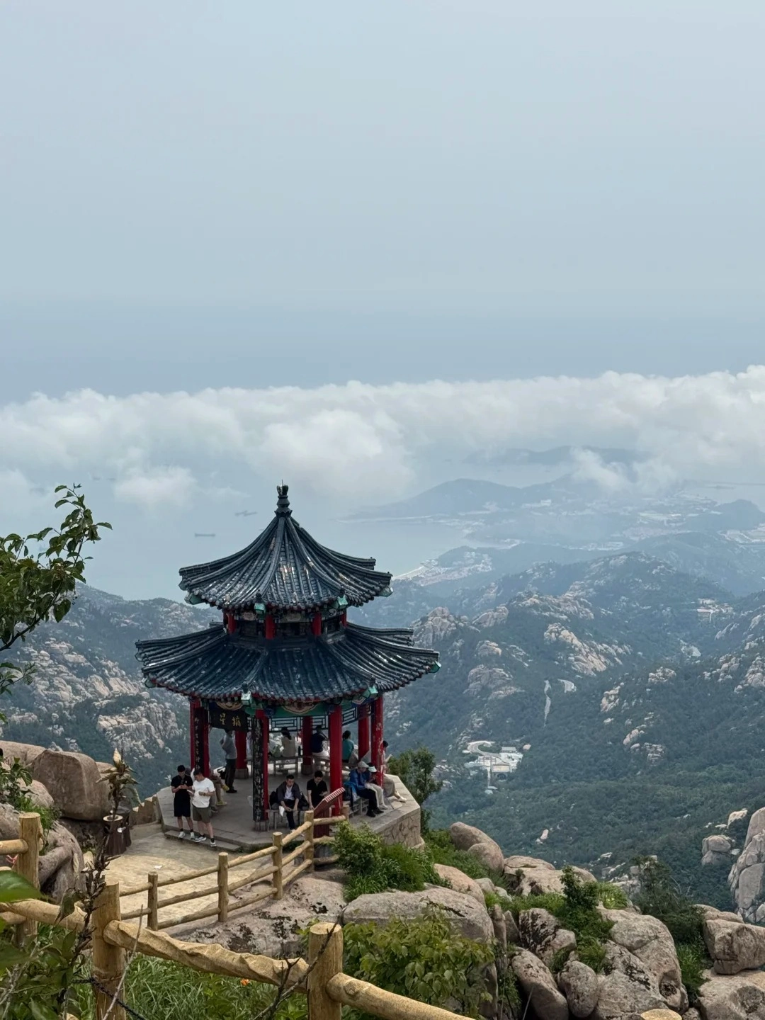A traditional Chinese pavilion with a dark blue tiled roof and red pillars sits on a rocky outcrop overlooking a vast landscape of mountains, green forests, and a cloudy expanse above the Yellow Sea.