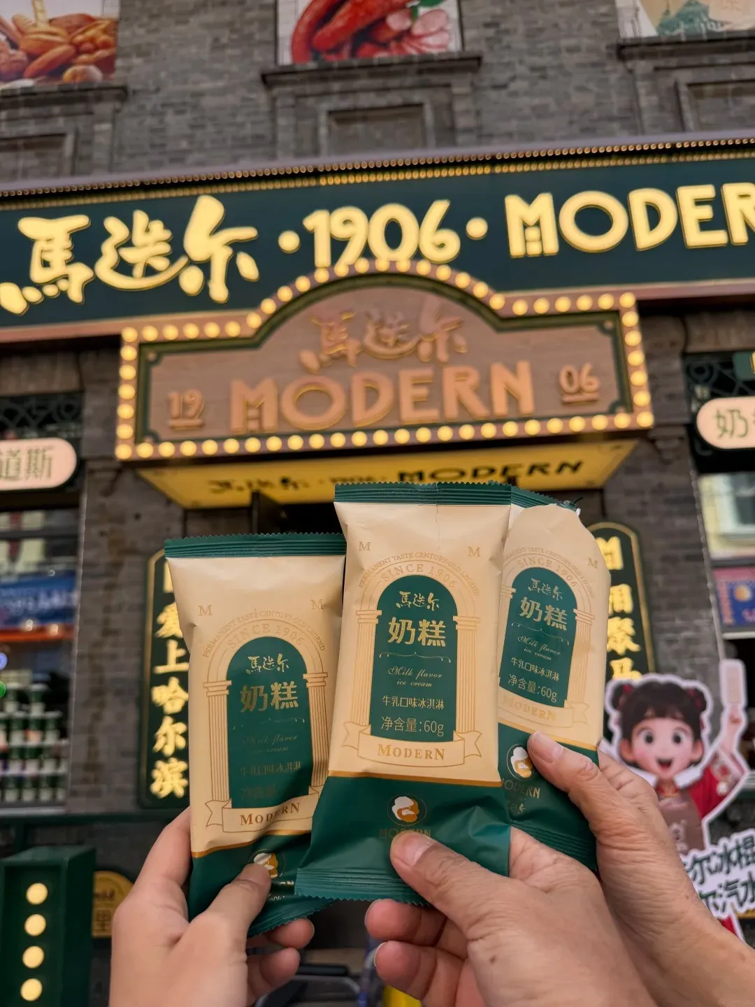 Two hands hold three packaged Madier milk flavor ice cream bars in front of a building with a sign reading '馬迭爾·1906·MODERN' and '馬迭爾 MODERN 06'.