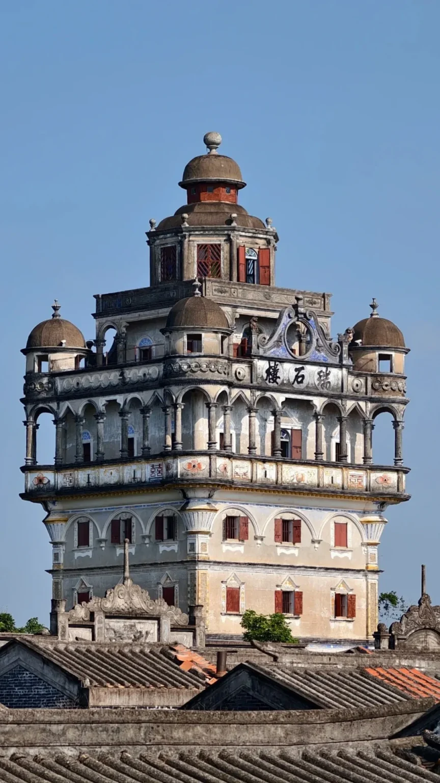 A tall, multi-story watchtower with a base of traditional Chinese tiled roofs, extending upwards with multiple levels of balconies, arches, columns, and domes. The tower is primarily light-colored with decorative bands and red-shuttered windows. The top features a prominent central dome and smaller flanking domes. Chinese characters are visible on an ornate section of the upper facade. The structure is set against a clear blue sky.