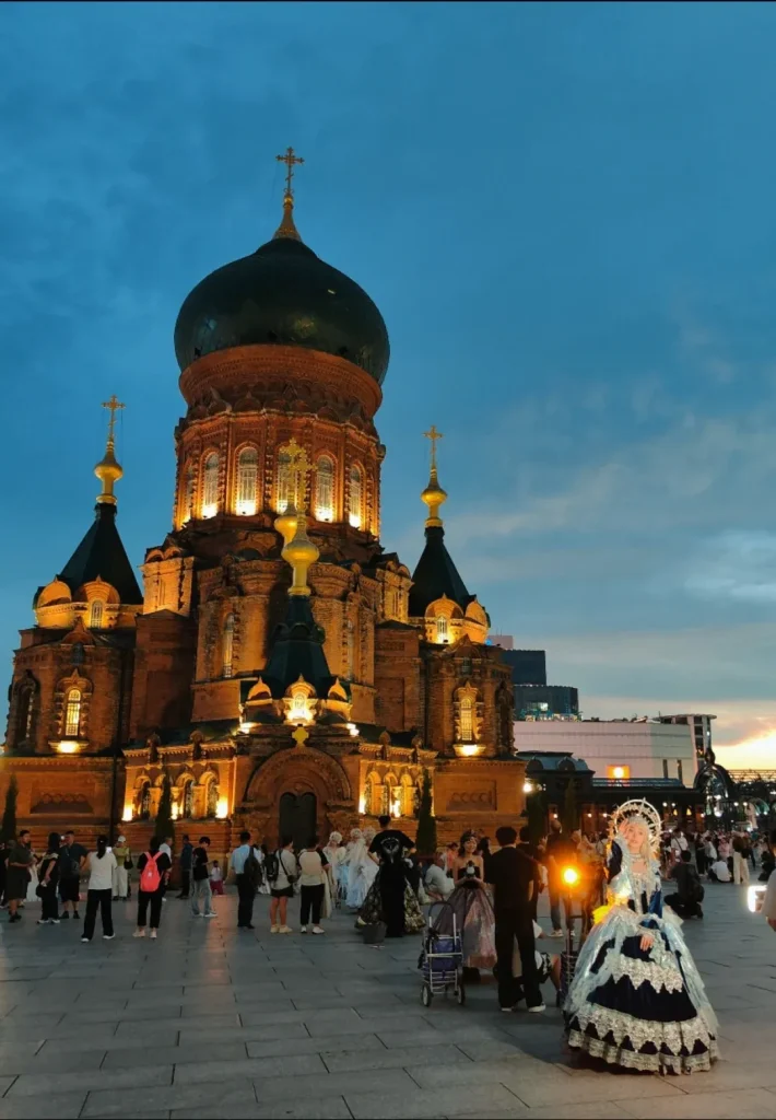 A large, ornate Russian Orthodox cathedral with a dark green dome and multiple gold crosses is illuminated at dusk. People are gathered in the plaza in front of the cathedral, some in elaborate costumes, and a woman in a blue and white Victorian-style dress stands prominently in the foreground.