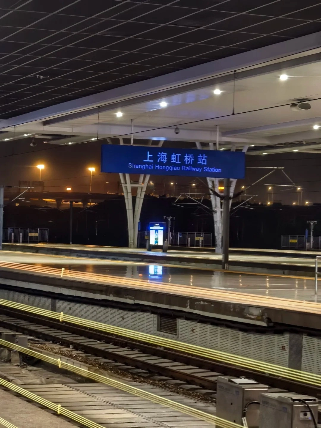 A nighttime view of an illuminated railway platform at Shanghai Hongqiao Railway Station. A blue sign displaying Chinese characters and 'Shanghai Hongqiao Railway Station' in English hangs overhead. Train tracks run in the foreground, with blurred streaks of light indicating movement. The platform itself is wet and reflects the overhead lights.